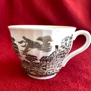 May include: White teacup with a brown landscape design. The cup has a handle and a detailed illustration of trees, buildings, and clouds. The teacup is set against a red background.
