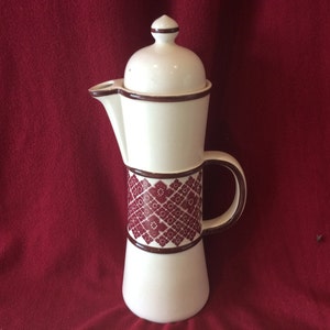 Peut inclure: Une cafetière en céramique blanche avec un motif géométrique bordeaux et brun. La cafetière a un couvercle et une poignée.