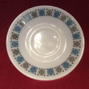 Può includere: Un piattino in ceramica bianca con un motivo geometrico blu e nero attorno al bordo. Il piattino ha un pozzetto centrale per una tazza.