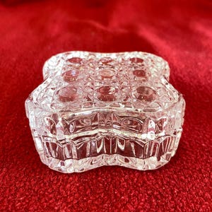 May include: A clear, cut-glass trinket box with a starburst pattern on the lid and sides. The box is shaped like a four-leaf clover and sits on a red surface. The box is likely used for jewelry or small keepsakes.