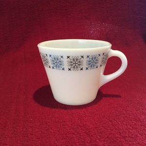 Puede incluir: Una taza de cerámica blanca con un patrón geométrico negro y azul. La taza tiene un asa y está sobre una superficie roja.