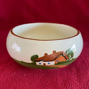 May include: A white ceramic bowl with a brown and green painted scene of a cottage with a brown roof and blue windows.