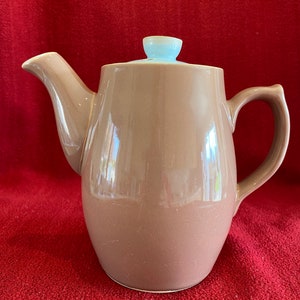 以下が含まれることがあります： 薄い青色の蓋が付いた茶色の陶器製のティーポット。ティーポットは丸みを帯びたボディで、片側に注ぎ口、反対側にハンドルが付いています。