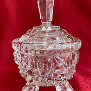 May include: A clear glass sugar bowl with a lid and three legs. The bowl is decorated with a geometric pattern.