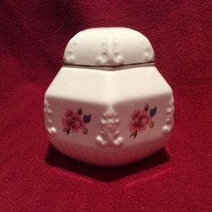 May include: White ceramic jar with a lid, featuring a floral design with pink roses and gold accents. The jar has a hexagonal shape and a raised, embossed design.