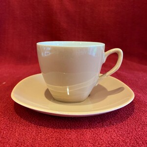 Puede incluir: Un juego de taza y platillo beige. La taza tiene un asa curvada y se asienta sobre un platillo a juego. El juego es de cerámica y de color neutro. El fondo es rojo liso.