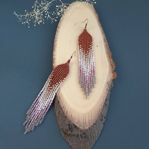 May include: A pair of long, beaded earrings with a gradient of colours, starting with copper at the top and fading to pink and grey at the bottom. The earrings are hanging from a wooden surface.