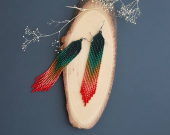 May include: A pair of long, beaded earrings with a gradient of red, orange, green, and black beads. The earrings are hanging from a wooden surface.