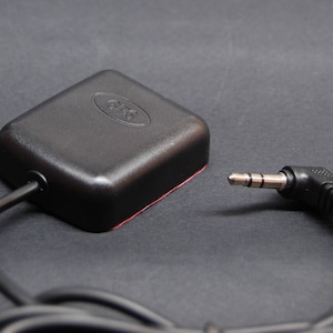 以下が含まれることがあります： 黒い正方形のGPSレシーバーで、黒いケーブルと3.5mmのオーディオジャックが付いています。レシーバーには「GPS」と書かれた小さなロゴがあります。
