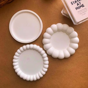 May include: Three white ceramic coasters. Two of the coasters have a raised, textured edge. The third coaster is smooth and flat.