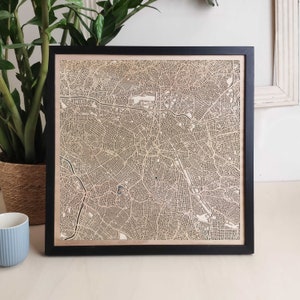 Op de afbeelding: Een ingelijste lasergesneden houten kaart van een stad. De kaart is gedetailleerd en ingewikkeld, met de straten en gebouwen van de stad. De lijst is zwart en de kaart is lichtbruin.