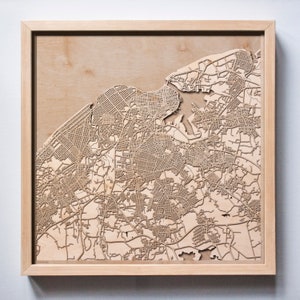 May include: A framed wooden map of a city with a detailed laser-cut design. The map is mounted on a light brown wood background and has a light brown wood frame.