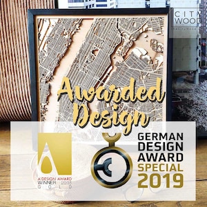 May include: A wooden map of a city with the text "Awarded Design" in gold lettering. The map is framed in black and has a gold award seal with the text "German Design Award Special 2019".