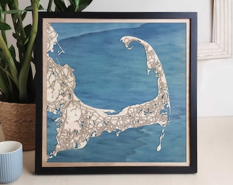 Op de afbeelding: Een houten kaart van Cape Cod, Massachusetts, met een blauwe en witte aquarel achtergrond. De kaart is ingelijst in zwart en is een 3D reliëfkaart.