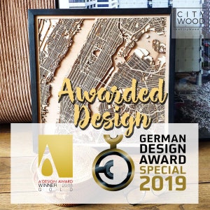 May include: Awarded Design, a laser-cut wooden map of a city, framed in black. The map is displayed on a wooden surface with two gold award logos. The text "A'DESIGN AWARD WINNER 2019 GOLD" is on the left logo. The text "GERMAN DESIGN AWARD SPECIAL 2019" is on the right logo.