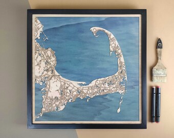Op de afbeelding: Een ingelijste houten kaart van Cape Cod, Massachusetts, met een blauwe en witte aquarel achtergrond. De kaart is gedetailleerd met wegen en steden. De lijst is zwart en de kaart is gemonteerd op een lichte houten achtergrond.