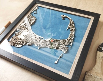 Op de afbeelding: Een ingelijste houten kaart van Cape Cod, Massachusetts, met een blauwe achtergrond en een gedetailleerde houten uitsnijding van de kustlijn. De kaart is zwart ingelijst en heeft een klein rood punt dat de locatie van Provincetown markeert.
