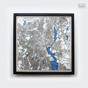 Pode incluir: Um mapa de madeira de Boston, Massachusetts, cortado a laser, com uma característica de água azul. O mapa é emoldurado em preto e montado em um fundo branco.
