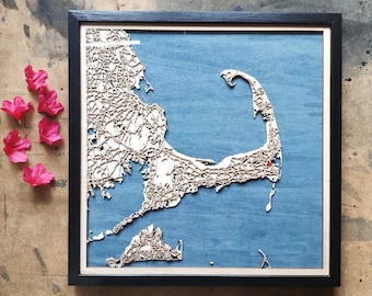 Op de afbeelding: Een houten kaart van Cape Cod, Massachusetts, met een blauwe achtergrond en een zwarte lijst. De kaart is gedetailleerd en toont de wegen en steden in het gebied. De kaart is een uniek en decoratief kunstwerk.