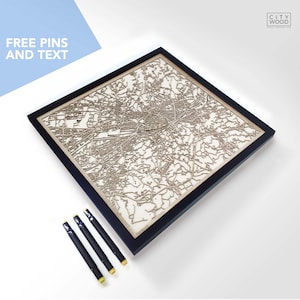 May include: A wooden map of a city in a black frame. The map is laser cut and shows the streets and landmarks of the city. The map is on a white background. The image also shows four black markers with yellow tips.