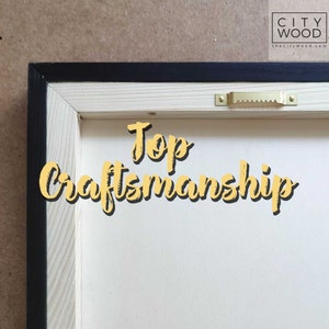 May include: Close-up of the back of a wooden frame with a black border. The frame is made of natural wood and has a gold-colored hanging hook. The text "Top Craftsmanship" is written in yellow on the frame.