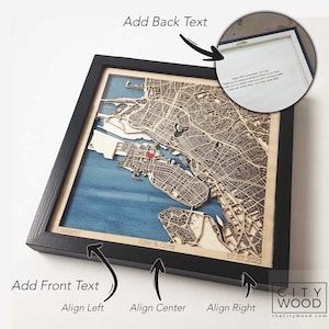 May include: A framed wooden map of Oakland, California with a blue background. The map is laser cut and has a 3D effect. The frame is black and the map is personalized with the text "Kate & Chris" and the date "07.07.2023".