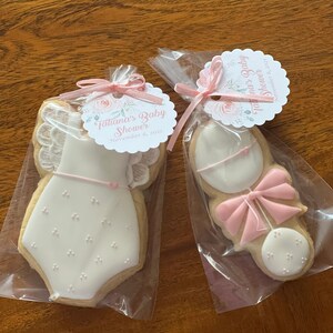 May include: Two decorated sugar cookies in clear cellophane bags. One cookie is shaped like a baby onesie with white icing and pink accents. The other cookie is shaped like a rattle with white icing and pink accents. Both cookies have a pink ribbon tied around them. The bags have tags with the text "Tatiana's Baby Shower" and the date "November 6, 2021".