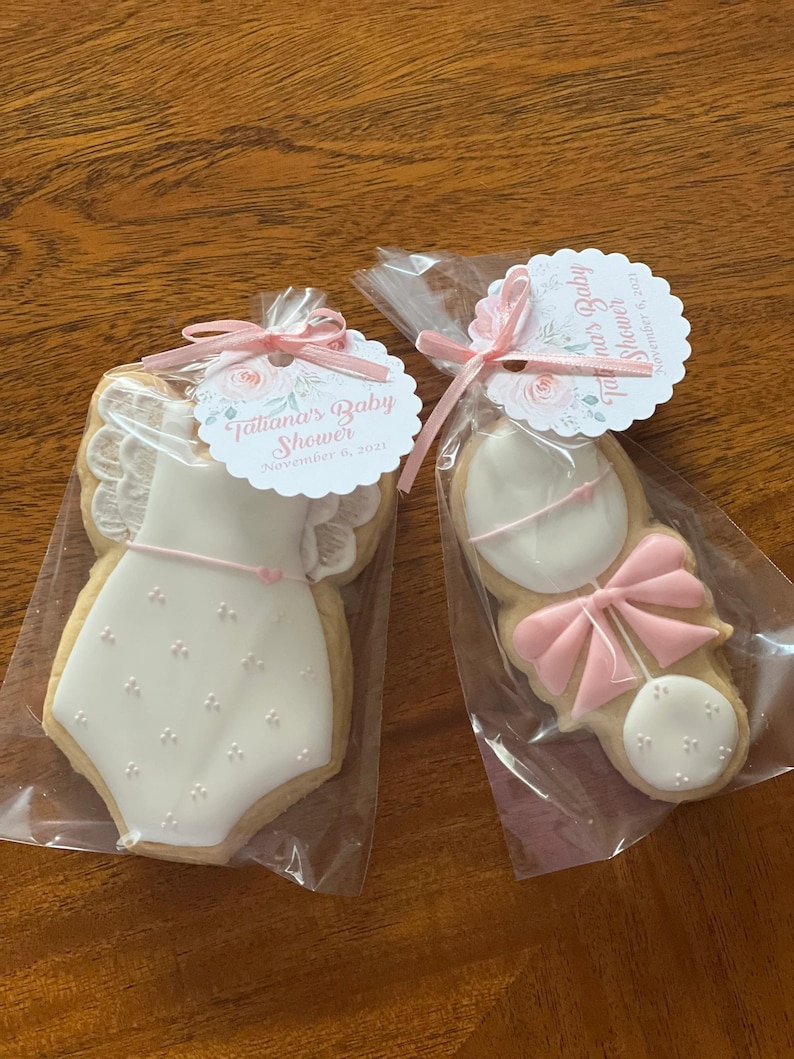 May include: Two decorated sugar cookies in clear cellophane bags. One cookie is shaped like a baby onesie with white icing and pink accents. The other cookie is shaped like a rattle with white icing and pink accents. Both cookies have a pink ribbon tied around them. The bags have tags with the text "Tatiana's Baby Shower" and the date "November 6, 2021".