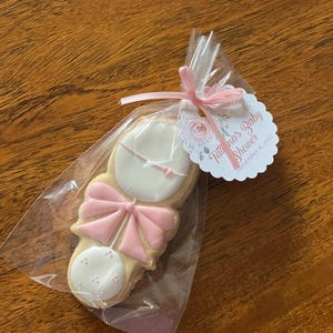 May include: A pink and white baby rattle cookie in a clear plastic bag with a pink ribbon. The tag attached to the bag reads "Tatiana's Baby Shower" and "November 6, 2021".