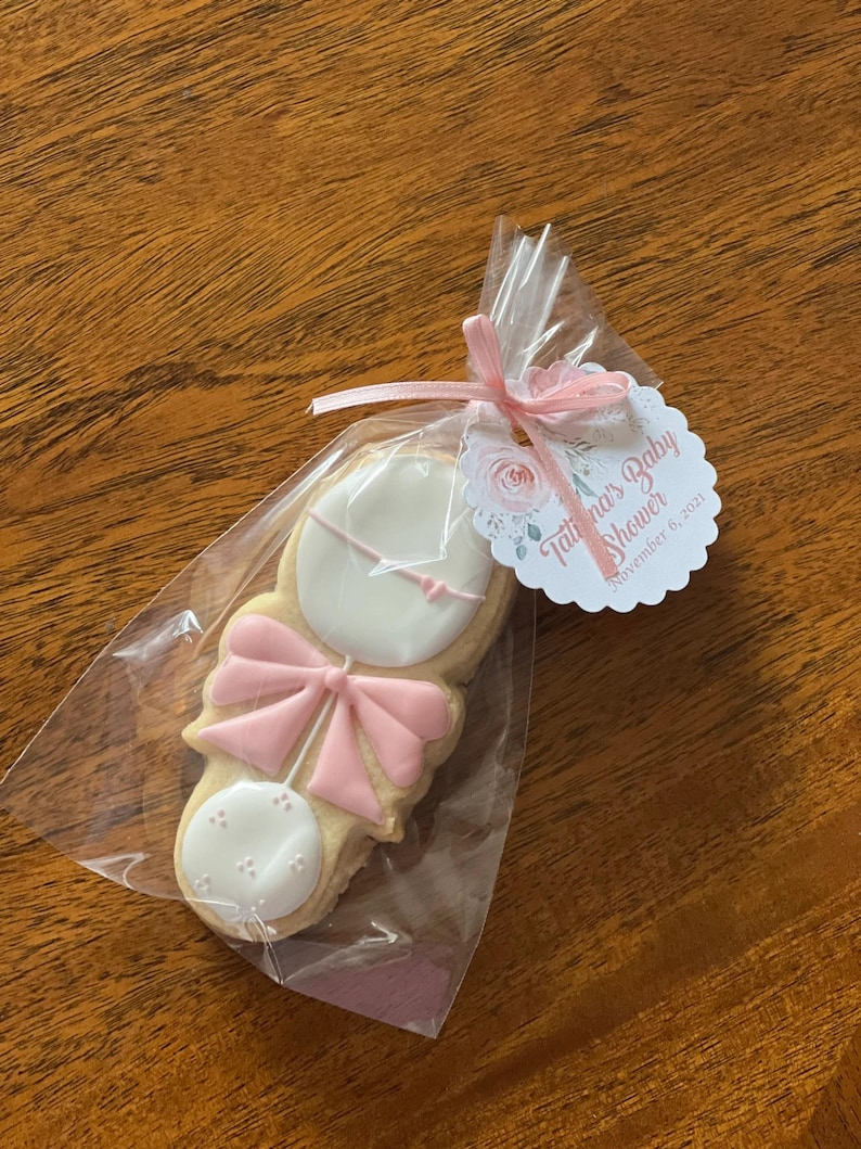 May include: A pink and white baby rattle cookie in a clear plastic bag with a pink ribbon. The tag attached to the bag reads "Tatiana's Baby Shower" and "November 6, 2021".