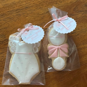 May include: Two decorated sugar cookies in individual cellophane bags. One cookie is shaped like a baby onesie with pink and white icing and the other is shaped like a rattle with pink and white icing. The bags are tied with pink ribbon and each has a tag that says "Fabulous Baby Shower" and "November 8, 2021".