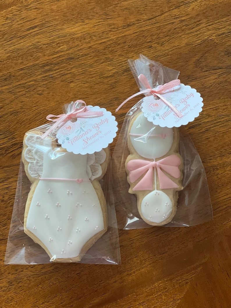 May include: Two decorated sugar cookies in individual cellophane bags. One cookie is shaped like a baby onesie with pink and white icing and the other is shaped like a rattle with pink and white icing. The bags are tied with pink ribbon and each has a tag that says "Fabulous Baby Shower" and "November 8, 2021".