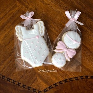 May include: Two decorated sugar cookies in individual cellophane bags tied with pink ribbon. One cookie is shaped like a baby onesie with pink polka dots and the other is shaped like a rattle with a pink bow.