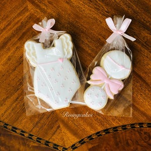 May include: Two decorated sugar cookies in cellophane bags. One cookie is shaped like a baby onesie with pink and white icing. The other cookie is shaped like a rattle with a pink bow and white icing.