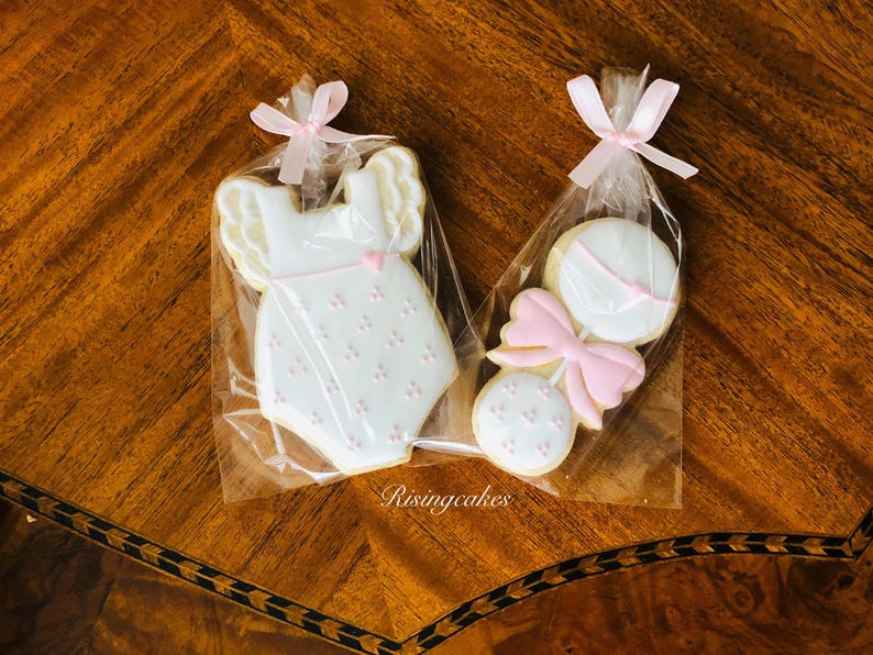 May include: Two decorated sugar cookies in cellophane bags. One cookie is shaped like a baby onesie with pink and white icing. The other cookie is shaped like a rattle with a pink bow and white icing.