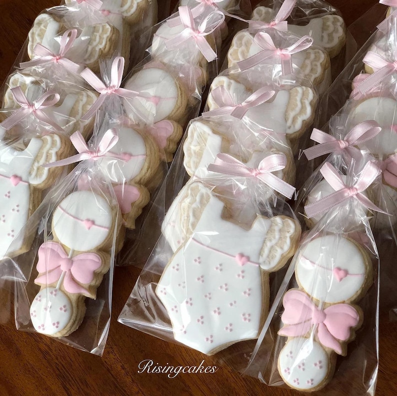 May include: A close-up of a set of baby shower cookies decorated with pink icing and white fondant. The cookies are packaged in clear cellophane bags with pink ribbons. The cookies are shaped like baby rattles and onesies.