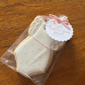 May include: A white cookie decorated with pink icing in the shape of a baby onesie. The cookie is packaged in a clear plastic bag with a pink and white tag that reads "Tatiana's Baby Shower November 6, 2021".