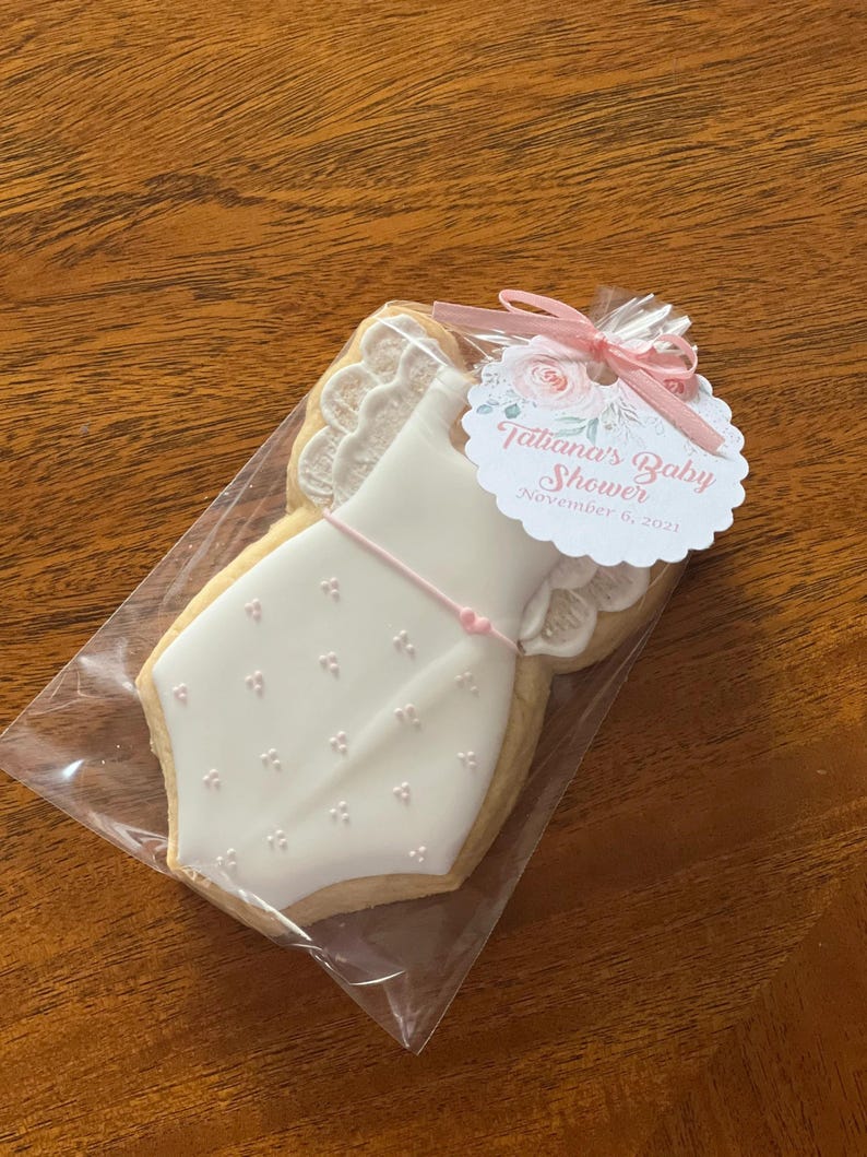 May include: A white cookie decorated with pink icing in the shape of a baby onesie. The cookie is packaged in a clear plastic bag with a pink and white tag that reads "Tatiana's Baby Shower November 6, 2021".