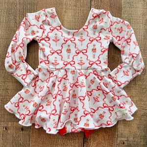 May include: A long-sleeve dress with a white base featuring a repeating pattern of red ribbons, nutcracker figures, and ballerinas. The garment has a scoop neckline, long sleeves with cuffs, and a flared skirt with a ruffled hemline.