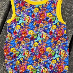 May include: A sleeveless romper featuring a vibrant pattern of Sesame Street characters in various colors. The romper has a yellow trim around the neckline and armholes, set against a dark purple background.