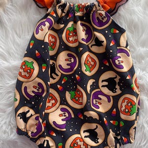 May include: A black romper featuring a repeating pattern of Halloween-themed cookies, including pumpkins, ghosts, and black cats, interspersed with candy corn. The garment has a gathered neckline and bright orange ruffled sleeves with black trim.