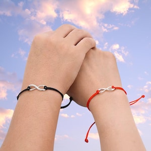 Könnte beinhalten: Zwei Armbänder mit Unendlichkeitszeichen-Anhängern. Eines hat eine schwarze Kordel, das andere eine rote Kordel. Die Unendlichkeitszeichen sind silberfarben. Die Armbänder befinden sich an Handgelenken vor blauem Himmel.
