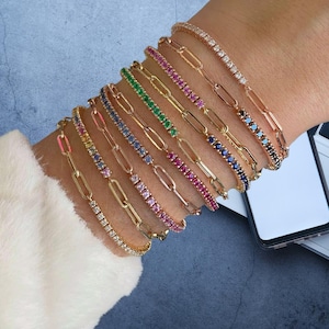 Puede incluir: Una colección de pulseras doradas con varios acentos de piedras preciosas, incluyendo diamantes, piedras rosas, verdes, azules y de arcoíris. Las pulseras se muestran en una muñeca, mostrando sus diferentes diseños y colores.