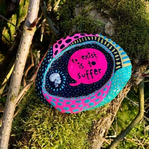 Könnte beinhalten: Ein bemalter Stein mit einem pinkfarbenen, blauen und schwarzen Design. Der Stein hat eine Sprechblase mit dem Text "to exist is to suffer" in weiß.