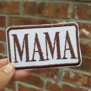 May include: A rectangular white patch with the word "MAMA" embroidered in brown capital letters. The patch has a brown border and is held against a brick wall background.
