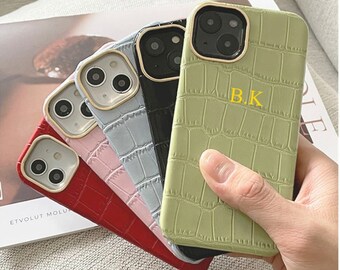 May include: Five different colored phone cases with a crocodile skin pattern. The cases are all black with gold trim around the camera lens. The case in the front has the initials "B.K." in gold lettering.