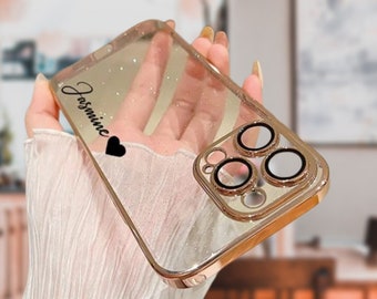 May include: A clear phone case with a gold trim and glitter. The case has a black heart and the name "Jasmine" written on it. The case has a raised camera lens protector with gold rings.