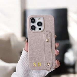 May include: A light beige leather phone case with a built-in finger grip and a gold button. The case has a textured surface and the initials "S.B." are embossed on the back.