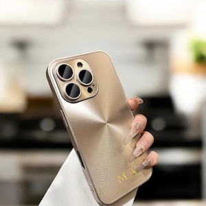 Puede incluir: Una funda de teléfono dorada con acabado de metal cepillado y un recorte para el objetivo de la cámara. La funda tiene las iniciales "MA" impresas en la parte posterior.