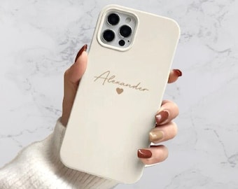 Puede incluir: Carcasa de teléfono blanca con una inscripción dorada que dice "Alexander" con un pequeño símbolo de corazón.